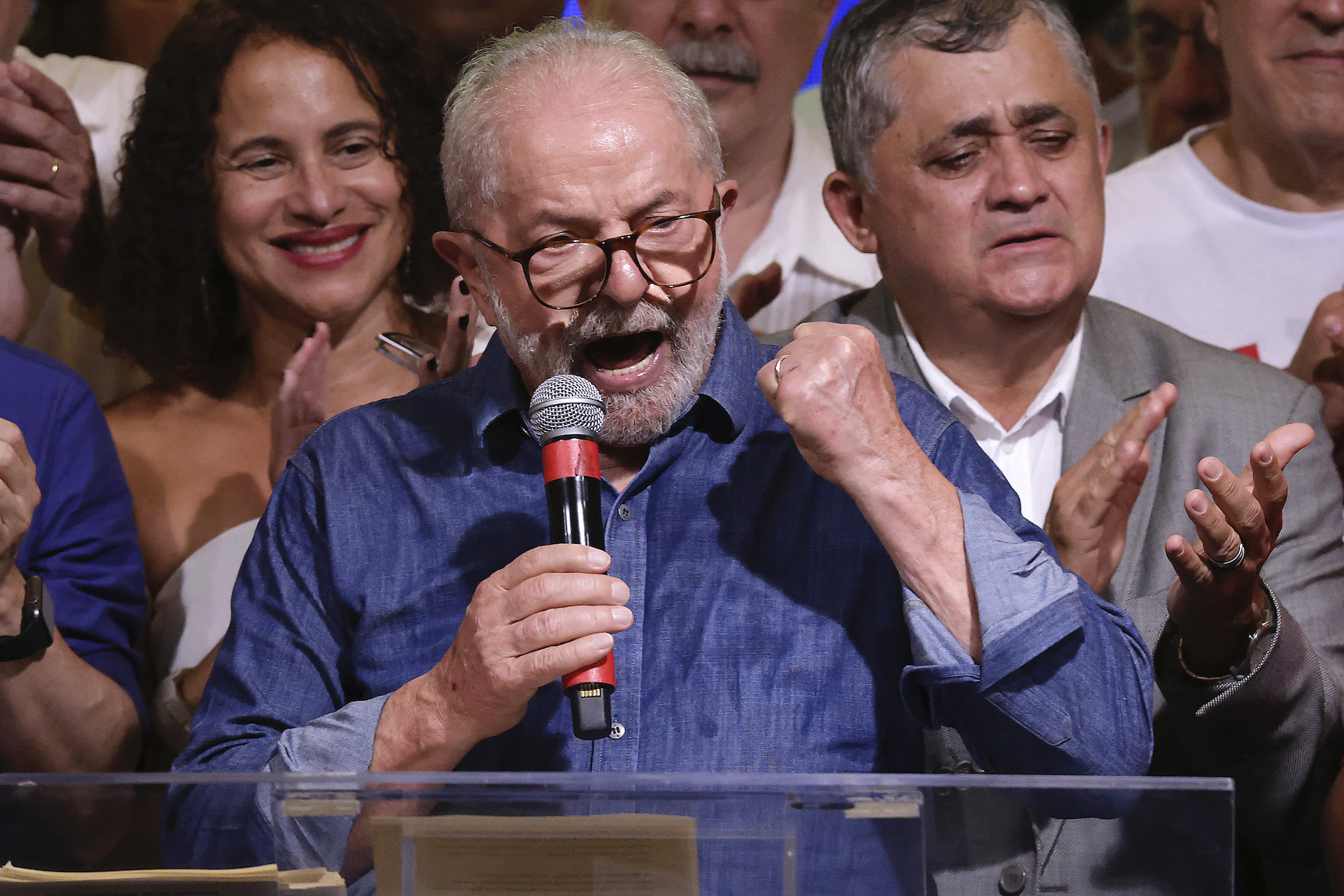 Lula holds a microphone and pumps his fist as he speaks. He stands behind a podium, and supporters are behind him. He wears a blue collared shirt and glasses.