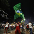 A man wearing a red Air Jordan jersey and shorts, red hat, and white glasses kneels in the middle of the street at night. He holds a Brazilian flag above his head with both hands. Around him are other people walking in the street, many of them also wearing red.