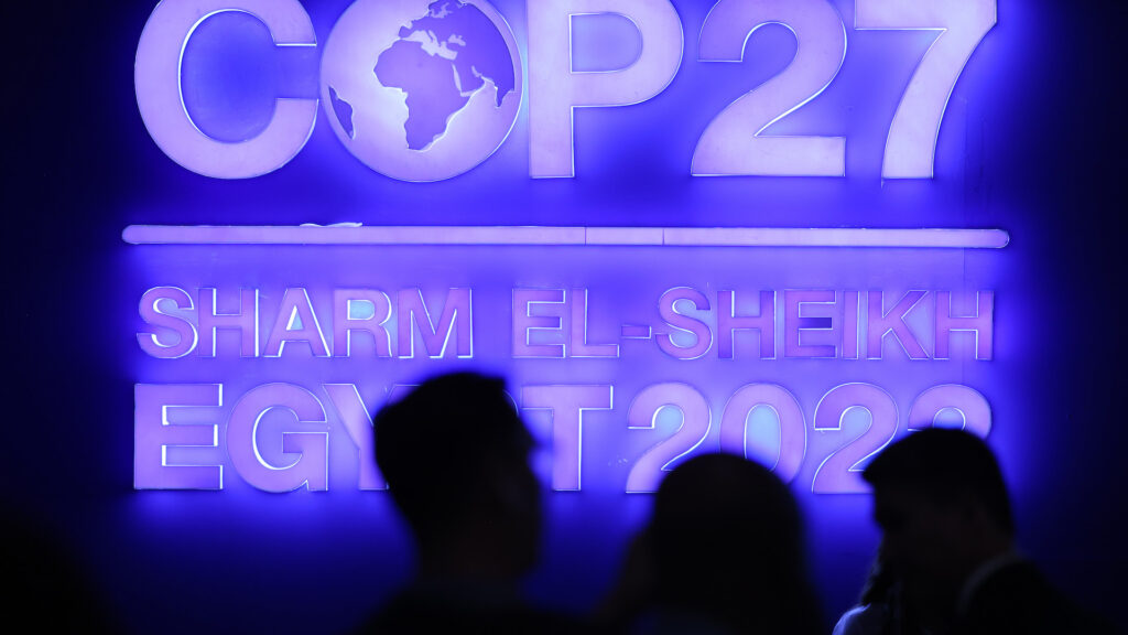 The silhouette of people's heads is in the foreground, illuminated by a large, purple neon sign in the background. The sign says COP27, Sharm El Sheikh, Egypt.