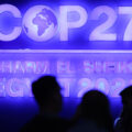 The silhouette of people's heads is in the foreground, illuminated by a large, purple neon sign in the background. The sign says COP27, Sharm El Sheikh, Egypt.
