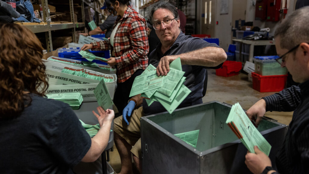 A man wearing glasses holds a pile of green envelopes in his hand and puts them in a net basket. Other workers around him also sort piles of green envelopes in what looks like a warehouse.