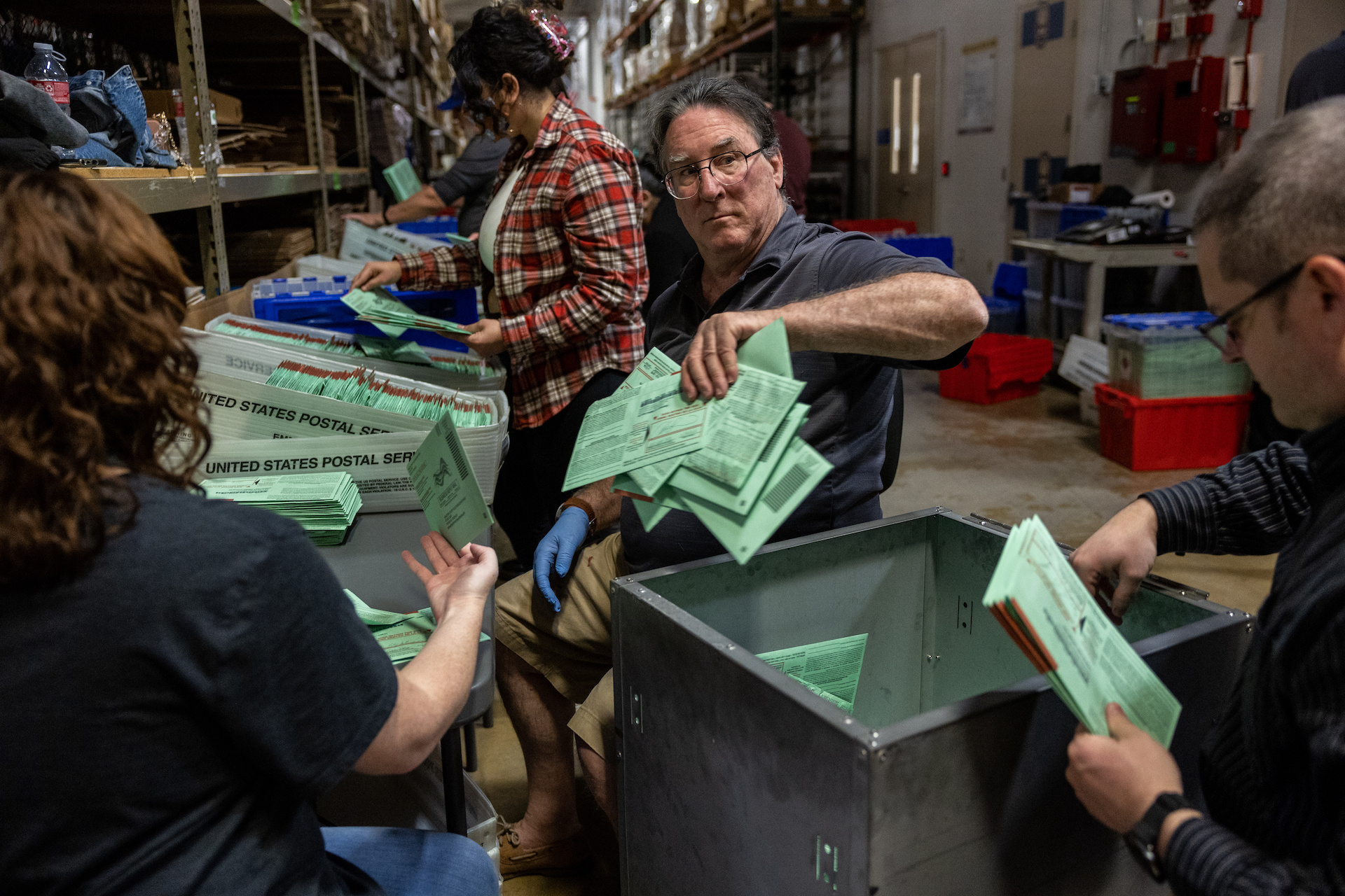 A man wearing glasses holds a pile of green envelopes in his hand and puts them in a net basket. Other workers around him also sort piles of green envelopes in what looks like a warehouse.
