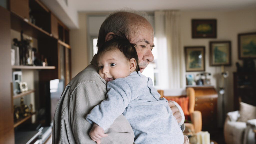 An elderly man holds a smiling infant in his arms. They are standing in a living room.