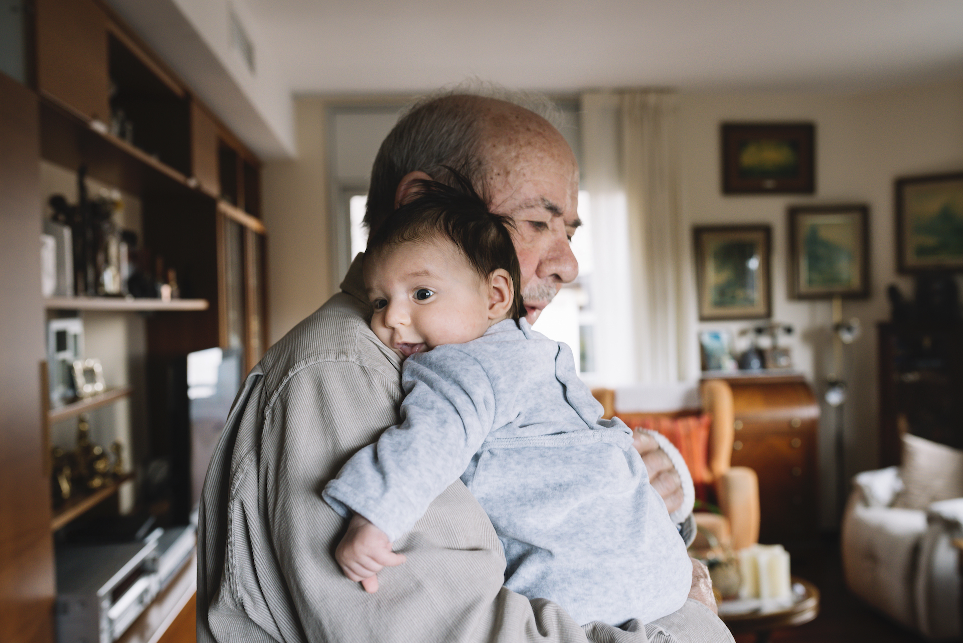 An elderly man holds a smiling infant in his arms. They are standing in a living room.