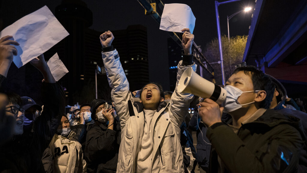A crowd of people stand outside at night. The central figure is a man holding both arms above his head and with is mouth open like he's shouting. In the right side of the photo, a man wearing a mask holds a megaphone to his mouth.
