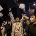 A crowd of people stand outside at night. The central figure is a man holding both arms above his head and with is mouth open like he's shouting. In the right side of the photo, a man wearing a mask holds a megaphone to his mouth.