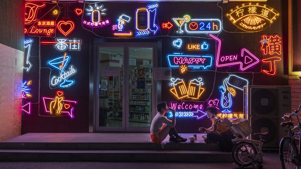 Two young women, one wearing a face mask, sit on the top step outside a closed store at night. The storefront is covered in colorful neon signs, which glow in the darkness.