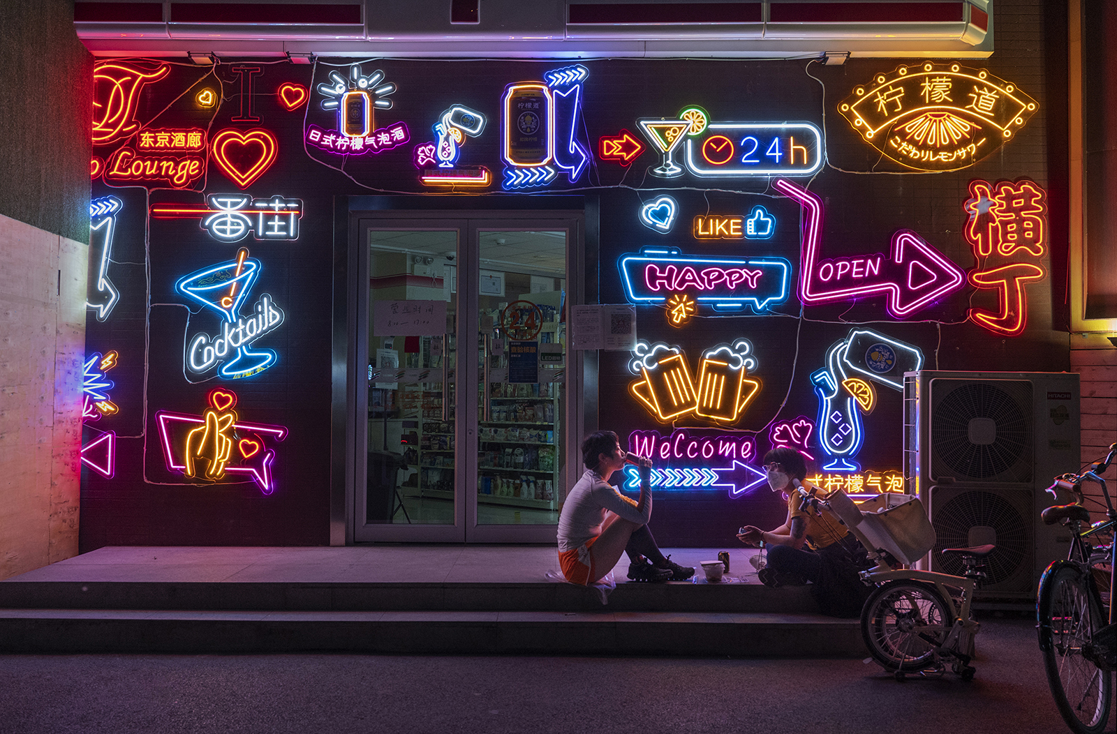 Two young women, one wearing a face mask, sit on the top step outside a closed store at night. The storefront is covered in colorful neon signs, which glow in the darkness.