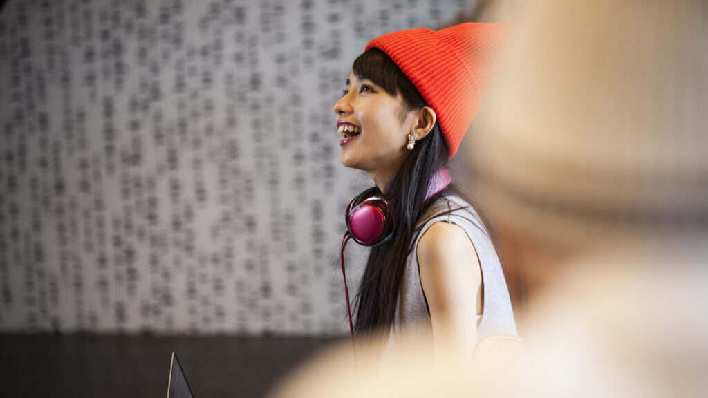 A young woman wearing an orange winter hat and pink headphones around her neck looks up and laughs.