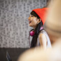 A young woman wearing an orange winter hat and pink headphones around her neck looks up and laughs.