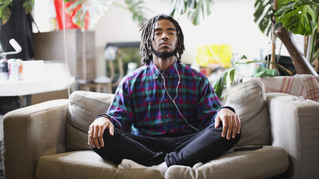 A man sits cross-legged on a couch while listening to music on white wired headphones. There are plants in the room behind him.