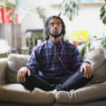 A man sits cross-legged on a couch while listening to music on white wired headphones. There are plants in the room behind him.