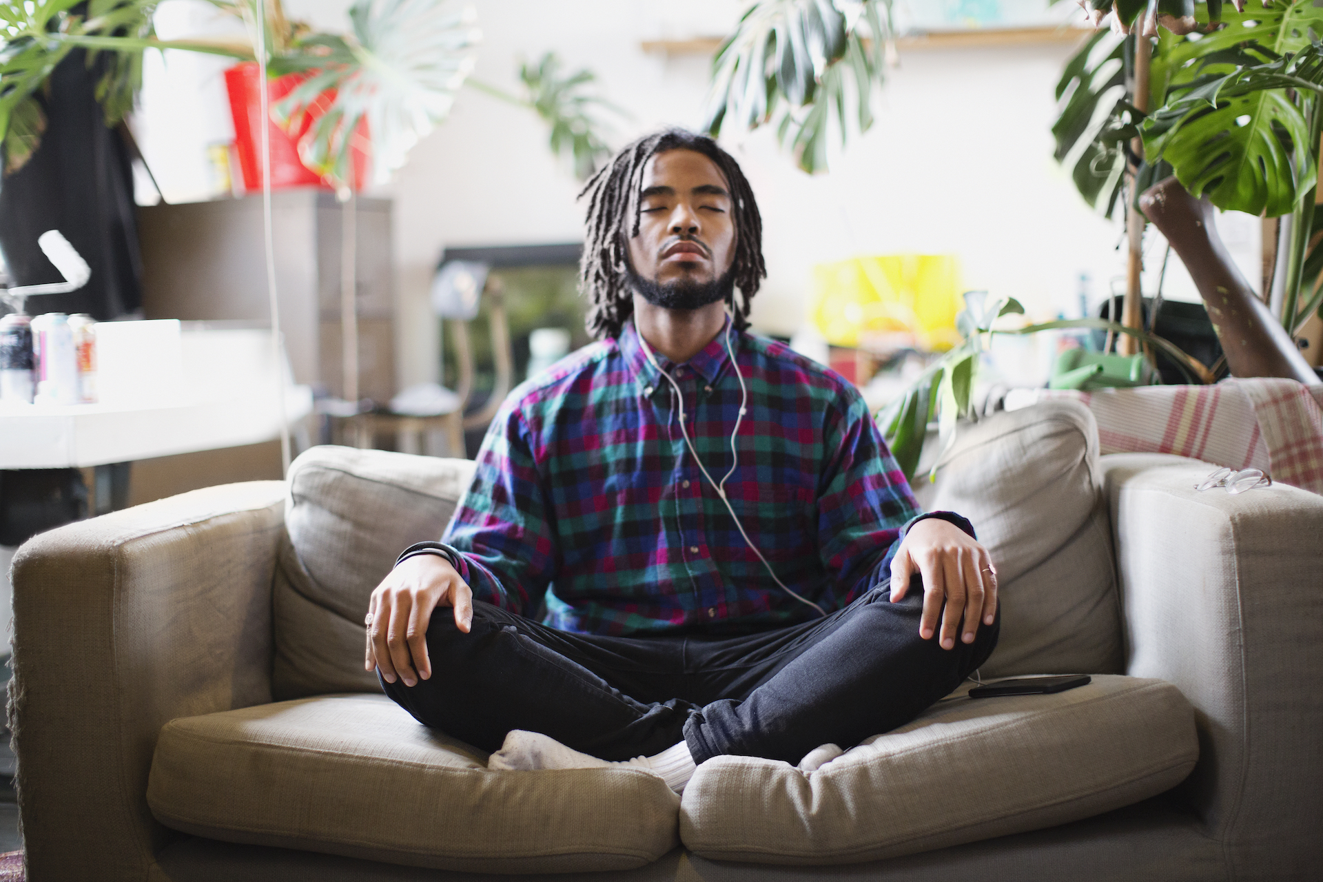 A man sits cross-legged on a couch while listening to music on white wired headphones. There are plants in the room behind him.