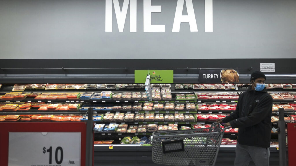 A man pushing a grocery cart stands in front of the meat section of a grocery store.