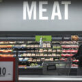 A man pushing a grocery cart stands in front of the meat section of a grocery store.