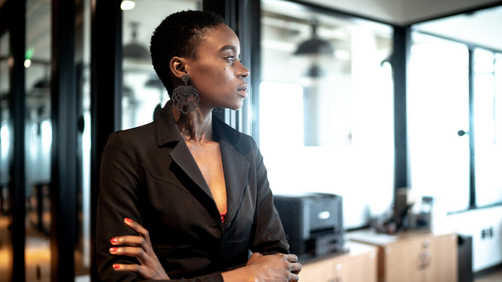 A young Black businesswoman in a black suit stands with her arms crossed. She is leaning against a glass door and looking out a window.