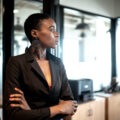 A young Black businesswoman in a black suit stands with her arms crossed. She is leaning against a glass door and looking out a window.