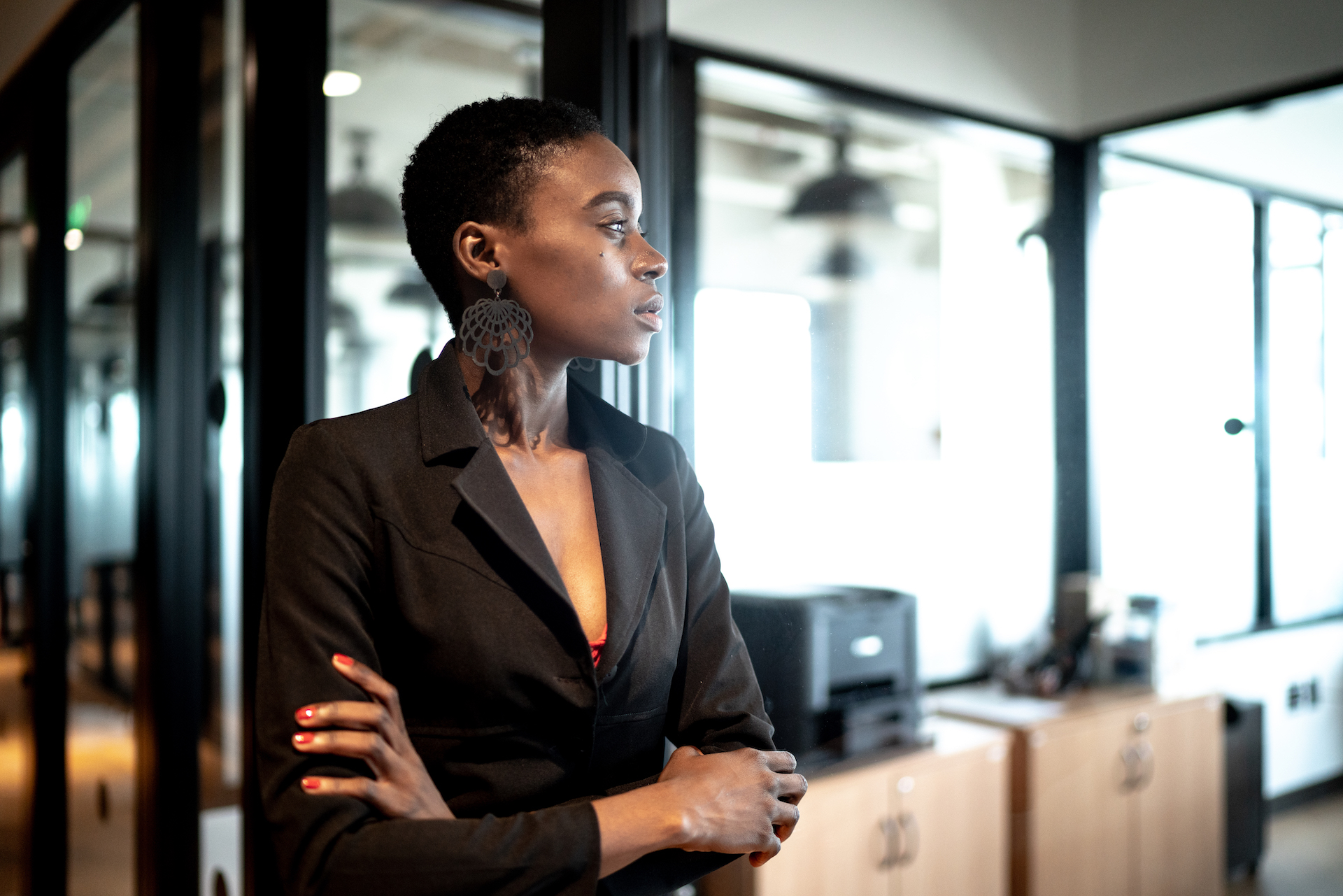 A young Black businesswoman in a black suit stands with her arms crossed. She is leaning against a glass door and looking out a window.