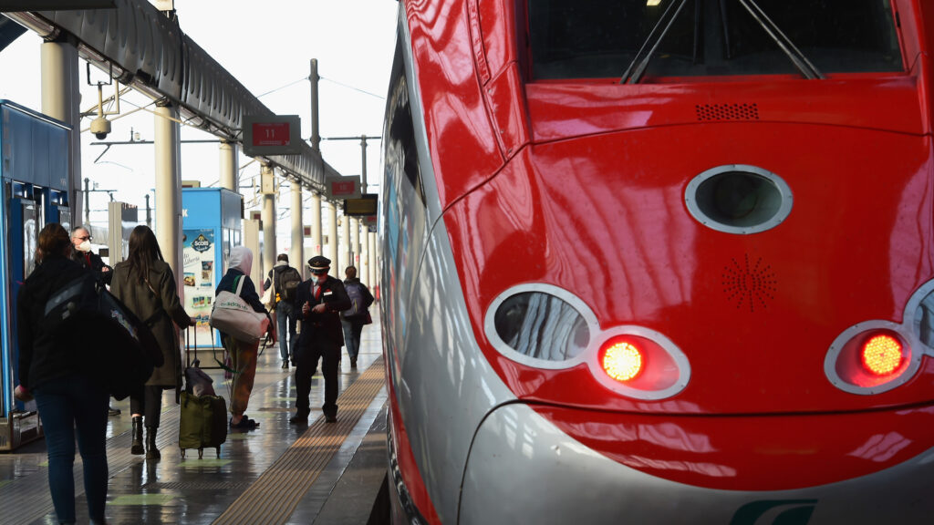 The nose of a high-speed train, which is painted a shiny red and grey. Passengers walk along the platform beside the railway.