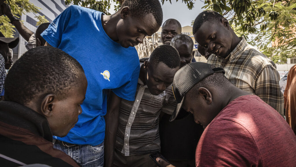 A group of men crowd around a sitting man, all of them looking at his cellphone. They are outside, and tree branches can be seen above them.