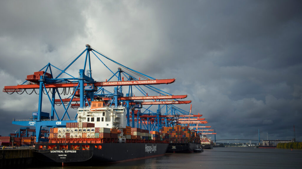 Ships filled with containers are docked in a harbor under a cloudy sky. Cranes rise overhead.