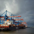 Ships filled with containers are docked in a harbor under a cloudy sky. Cranes rise overhead.
