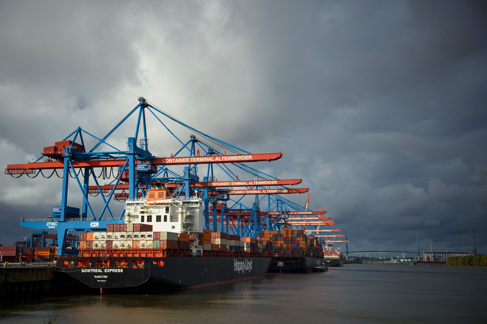 Ships filled with containers are docked in a harbor under a cloudy sky. Cranes rise overhead.