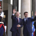 Two men in suits stand next to each other on the steps of a building, waving to a crowd. Two guards in blue and red ceremonial dress stand on either side of the doorway.