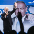 A man with grey hair wearing a dark suit stands on stage and raises one hand in the air while holding a microphone in the other. Behind him is a digital screen with his face on a campaign poster and the Israeli flag, a blue star of David on a white field. In front of him are the silhouettes of a crowd.