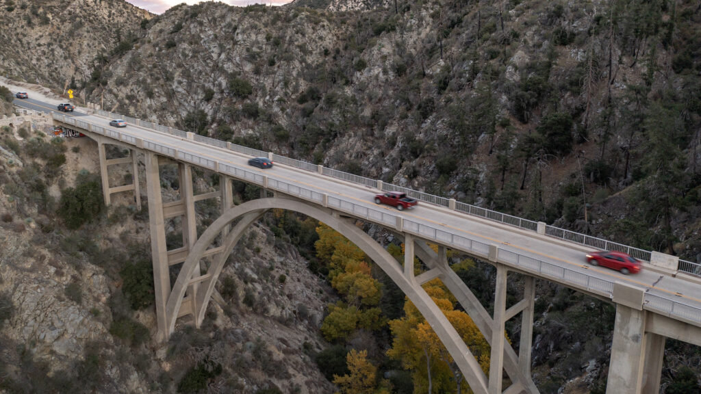 A bird's eye view of a bridge over a canyon, with cars on the road. In the middle of the canyon are trees turning yellow for fall.
