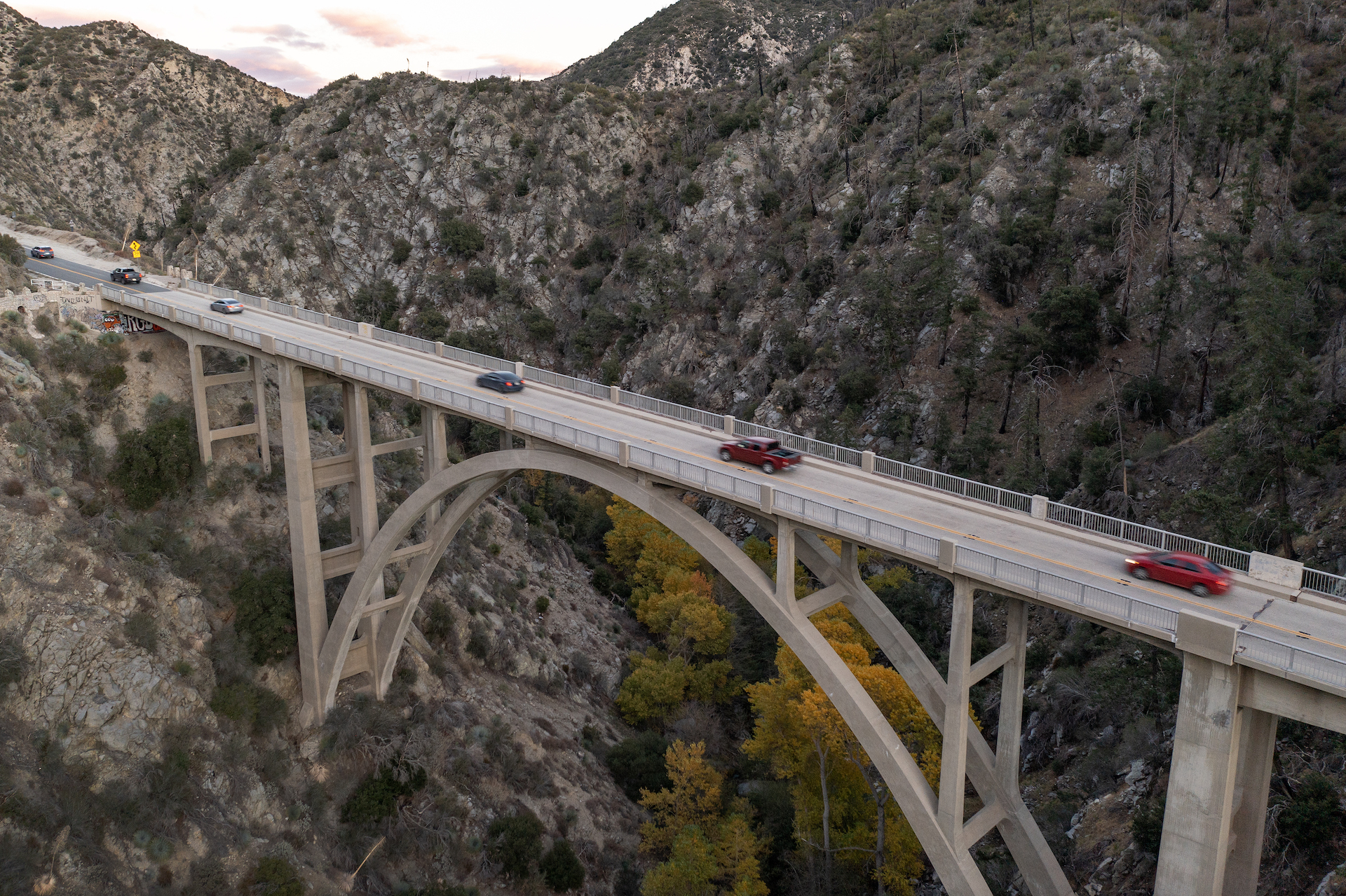A bird's eye view of a bridge over a canyon, with cars on the road. In the middle of the canyon are trees turning yellow for fall.