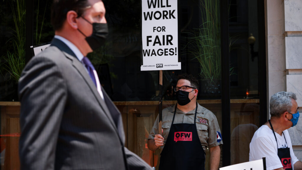A man wearing a black mask and apron stands on the sidewalk holding a sign that says, 