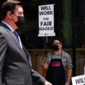 A man wearing a black mask and apron stands on the sidewalk holding a sign that says, 