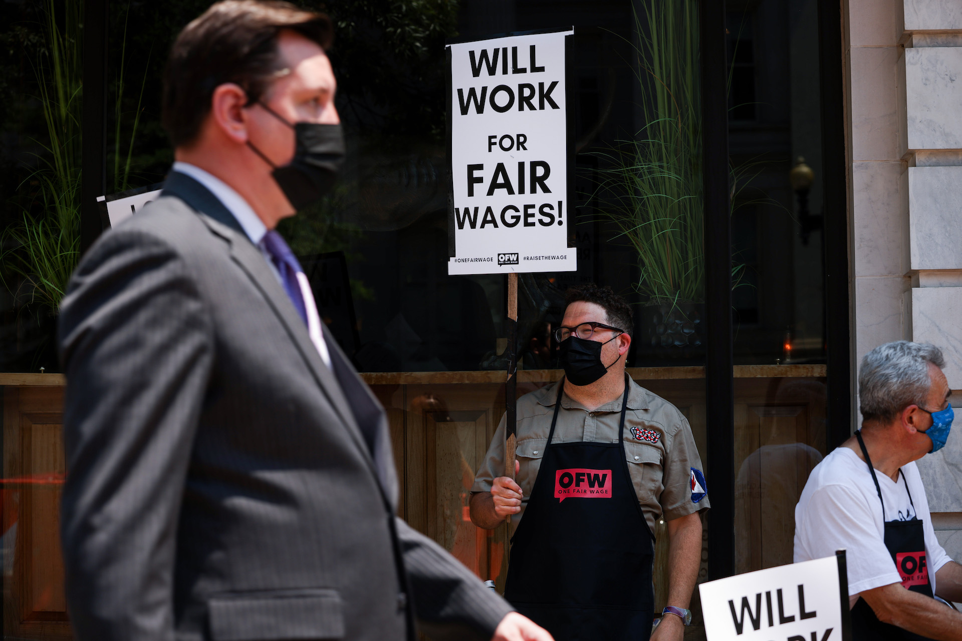 A man wearing a black mask and apron stands on the sidewalk holding a sign that says,