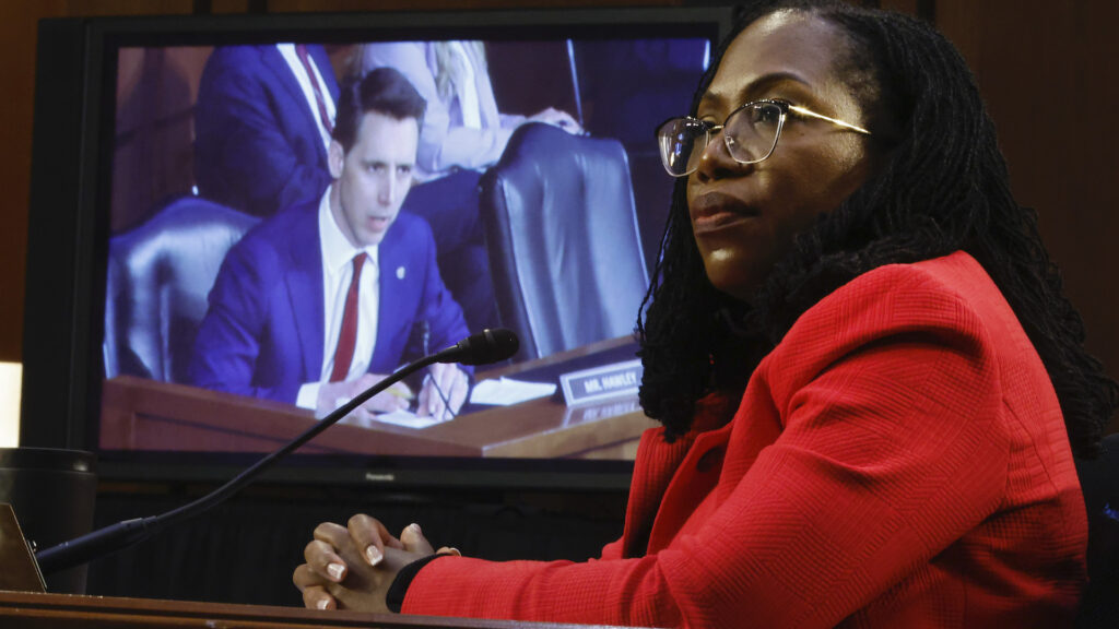 A Black woman in red jacket and glasses sits with her hands folded on a desk. Behind her is a screen showing a white man.