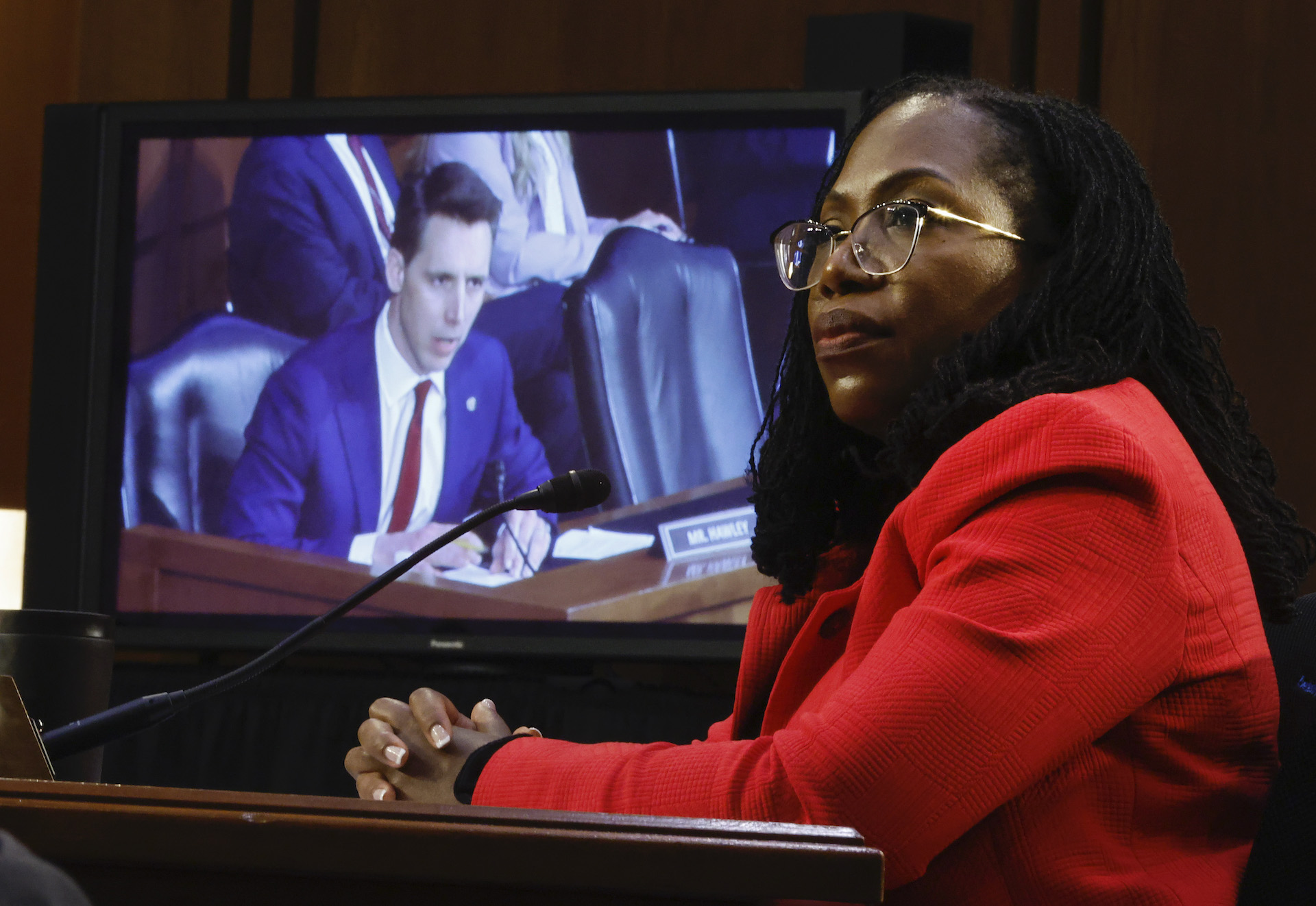 A Black woman in red jacket and glasses sits with her hands folded on a desk. Behind her is a screen showing a white man.