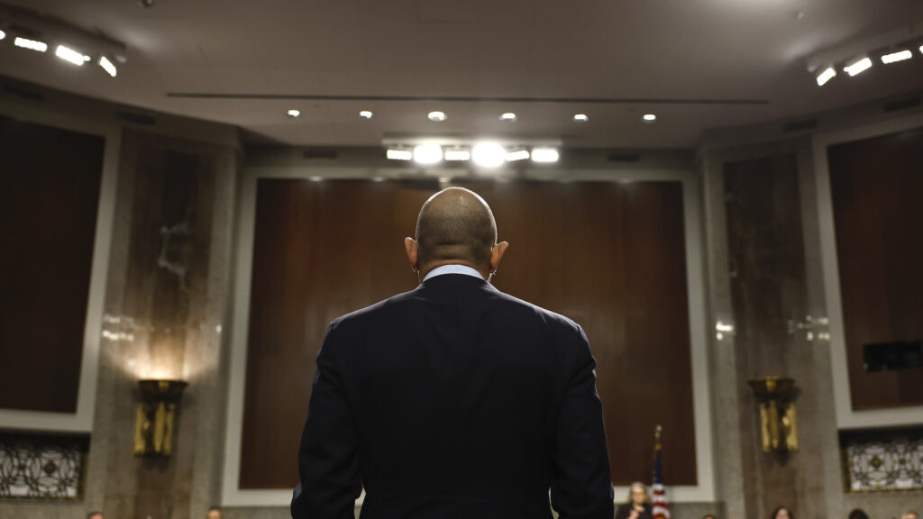 A man, seen from behind, stands in front of a large room, with a people sitting in the background. He is wearing a dark suit.