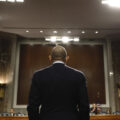 A man, seen from behind, stands in front of a large room, with a people sitting in the background. He is wearing a dark suit.