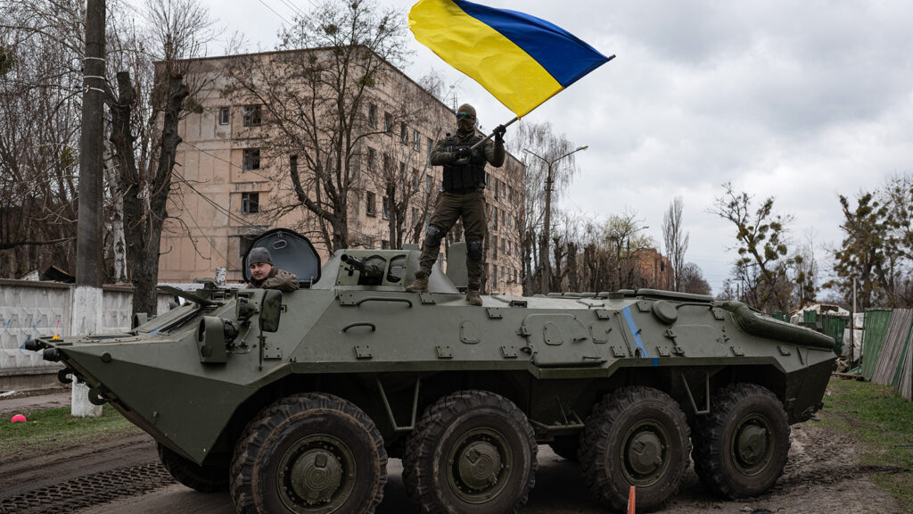 Two soldiers in camouflage are in a large green tank in the middle of a residential street. One soldier sits in the front of the tank. The other is standing on the vehicle, waving a blue and yellow Ukrainian flag.