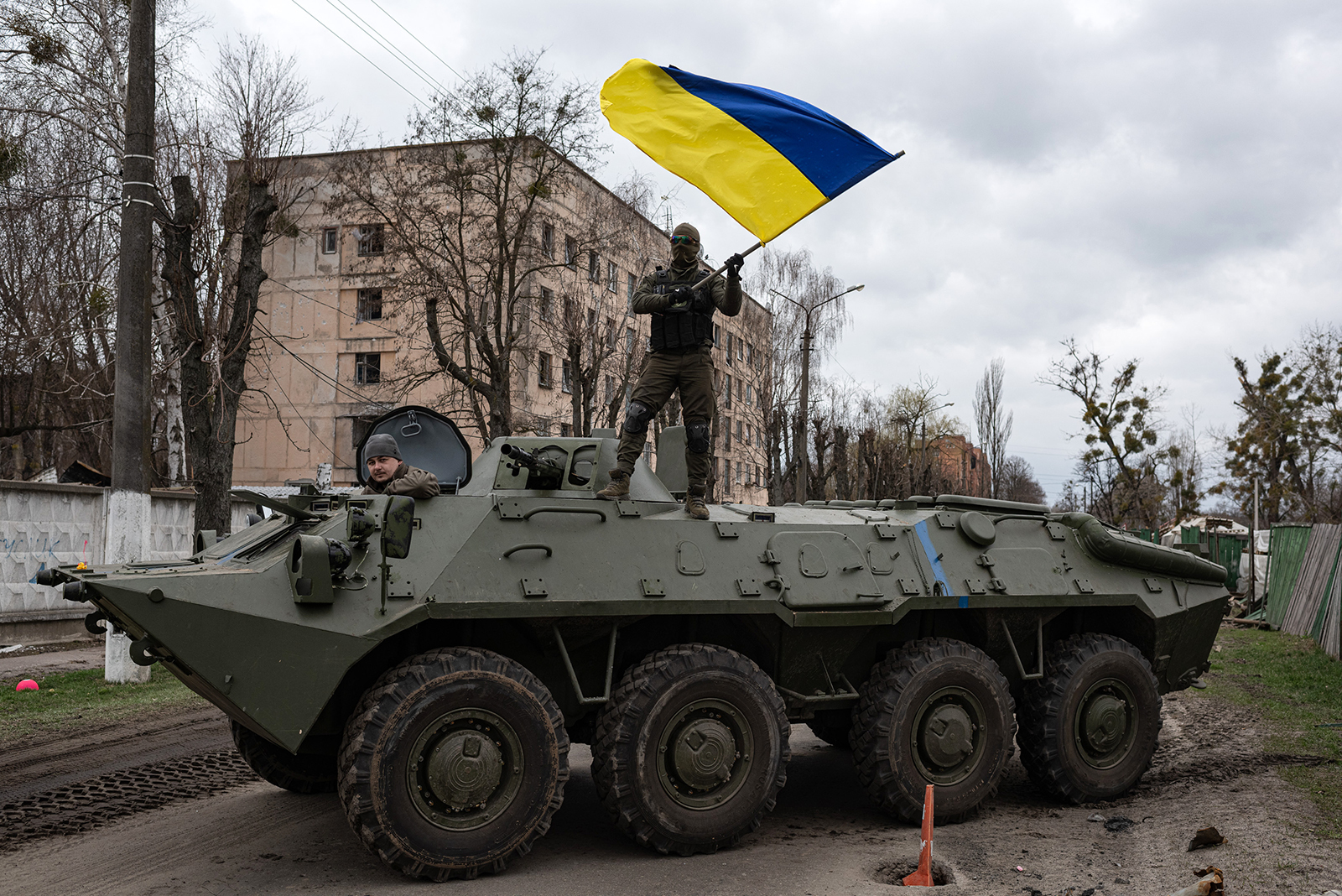 Two soldiers in camouflage are in a large green tank in the middle of a residential street. One soldier sits in the front of the tank. The other is standing on the vehicle, waving a blue and yellow Ukrainian flag.