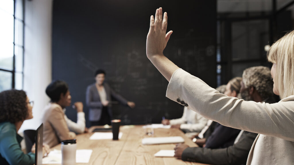 Businesswoman with hand raised sitting with male and female coworkers in conference room during office meeting